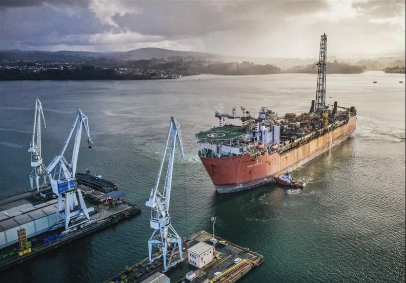  A red floating production storage and offloading unit is arriving at a dry dock in Ferrol, Spain. Three blue cranes on the dock tower over the vessel. The sun peers through the cloudy horizon where mountains are urban areas are seen behind the vessel.