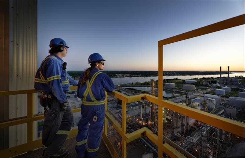 Two workers - Cori Senini and Kyle Ferguson - standing on a walkway at the top of a tower looking out at the Sarnia Refinery at sunrise. They are wearing blue hard hats, coveralls, safety glasses and gloves. The St. Clair River is in the background.