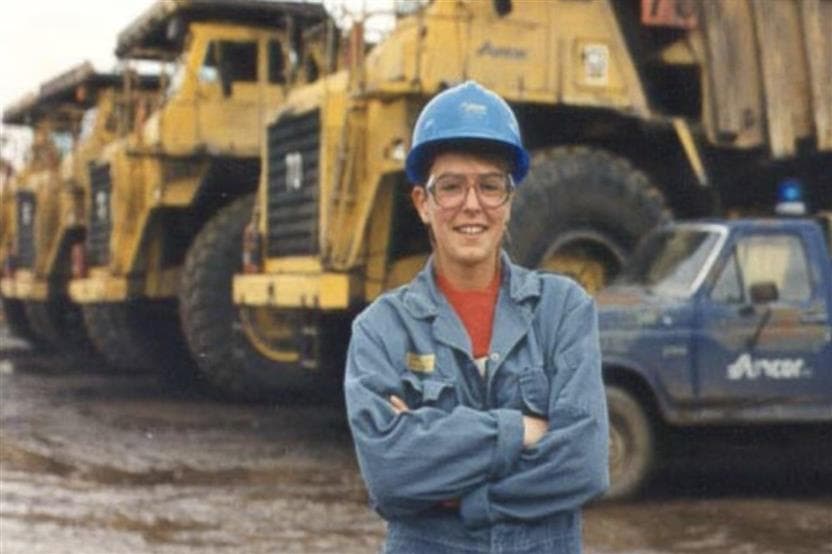 A female Suncor employee stands with her arms crossed in front of a line of Suncor equipment, one light vehicle and three haul trucks. She is wearing blue coveralls and a blue hard hat.