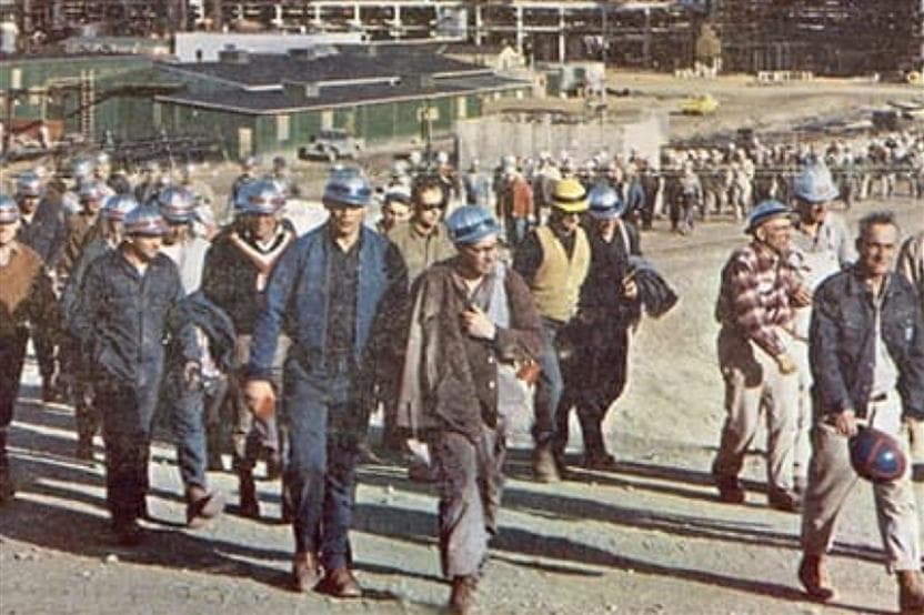 A large group of employees arrive for work at a Suncor site in the morning. Many are wearing blue hard hats and work clothes.