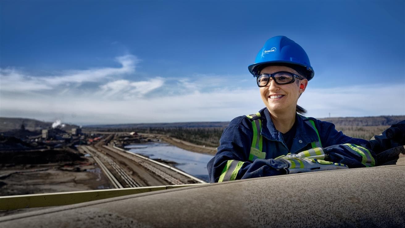 A woman in coveralls, PPE and a Suncor hard hat is smiling into the distance. There is blue sky and the Suncor operations in the background.